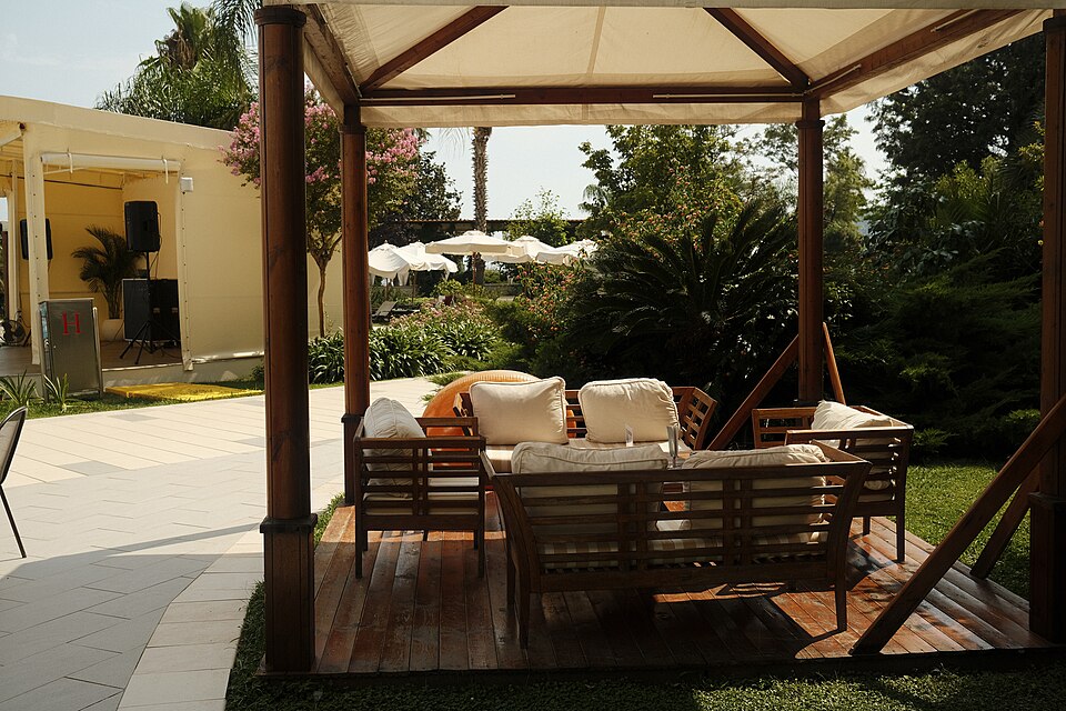 A cozy seating area featuring plush cushions and wooden framing sits in a vibrant garden. Surrounding greenery and sunlit pathways create a serene atmosphere.