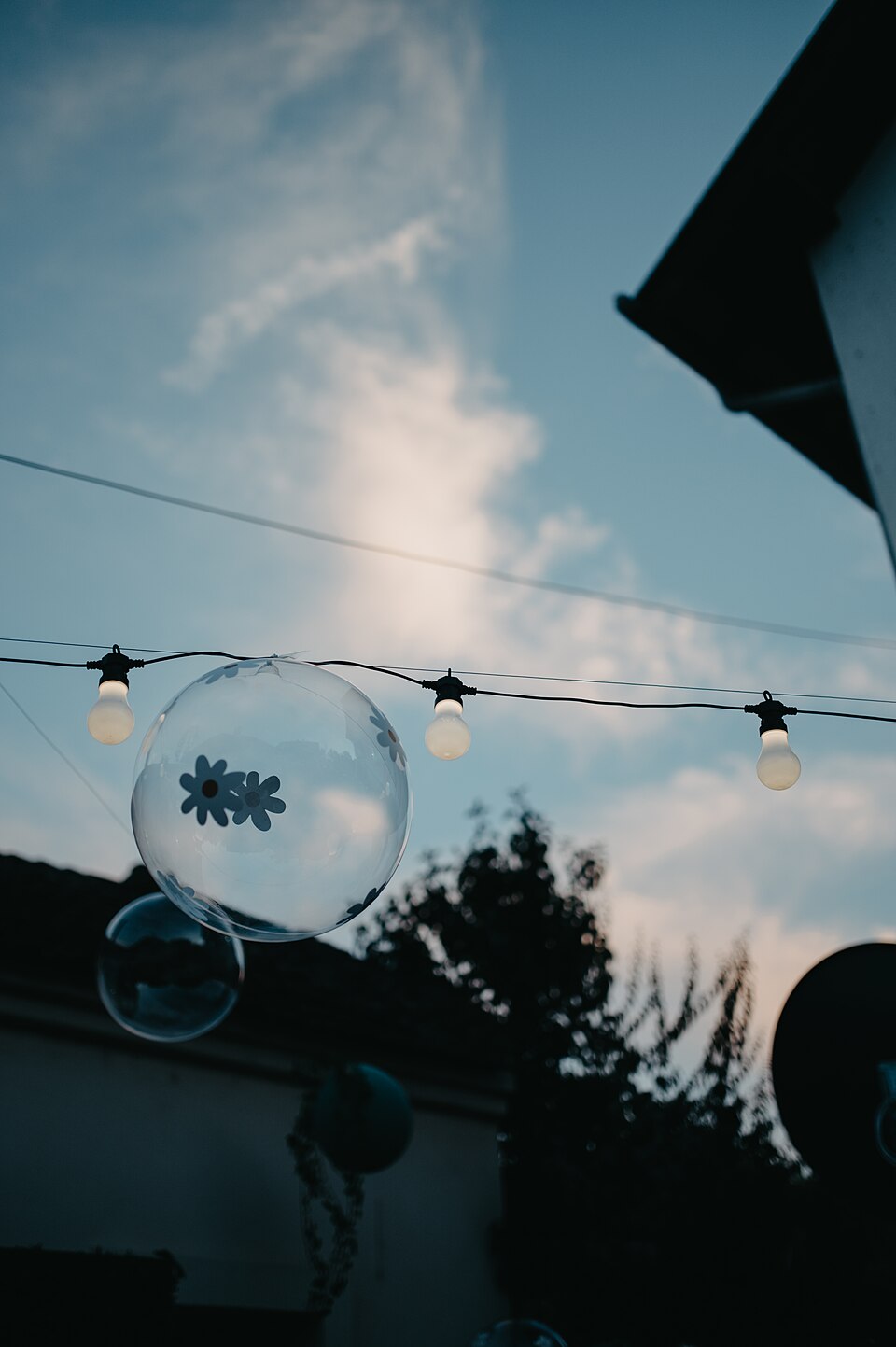 Colorful bubbles float gently in the air as strung lights illuminate a garden during dusk. The clouds reflect soft hues, adding to the tranquil ambiance.